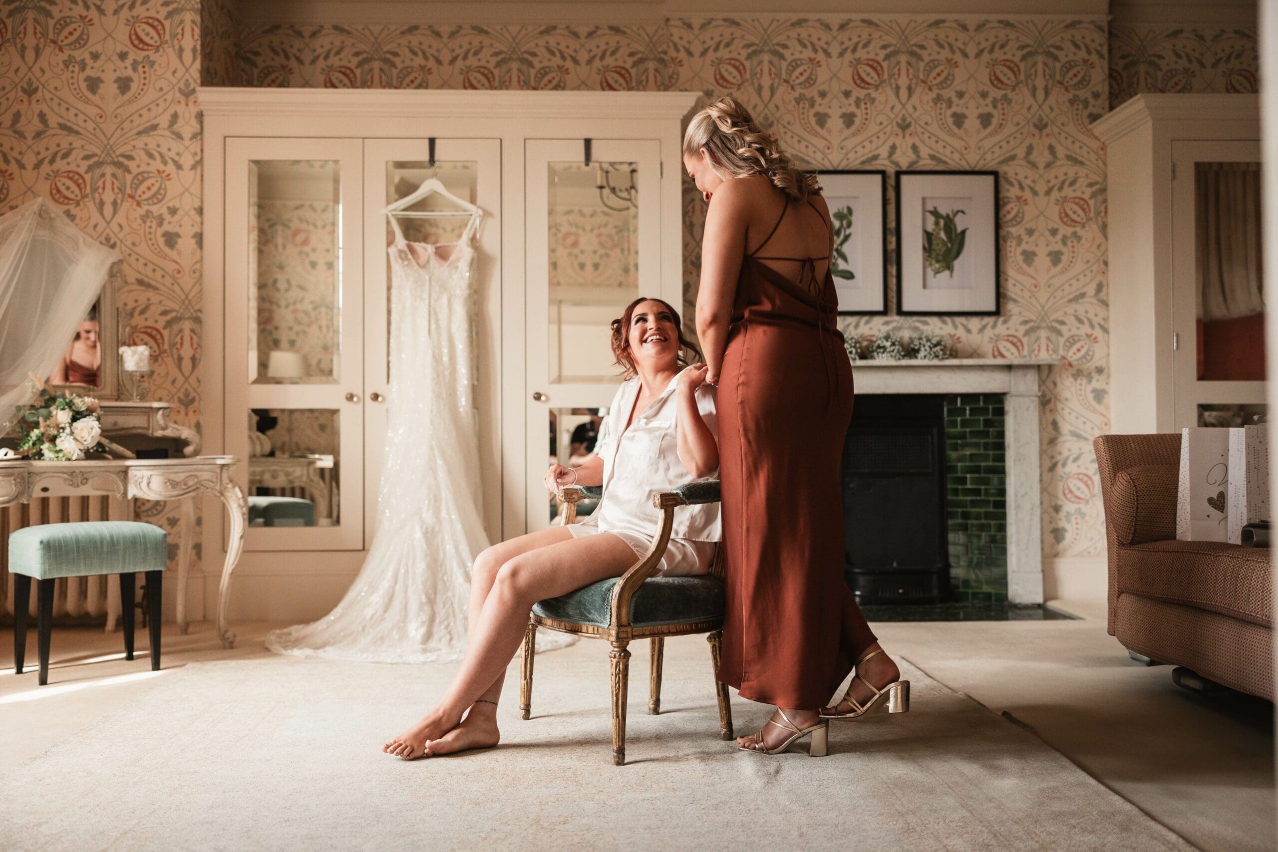 Bridesmaid dressed in bronze bridesmaid dress looking at her sister in bridal prep at hodsock priory before her wedding with the dress hanging in the background | Hodsock priory wedding photographer